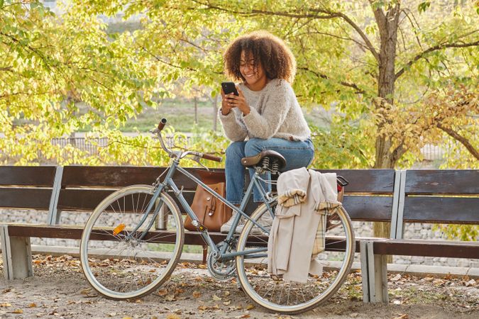 Smiling Black Latina woman resting on bench with smartphone near bike