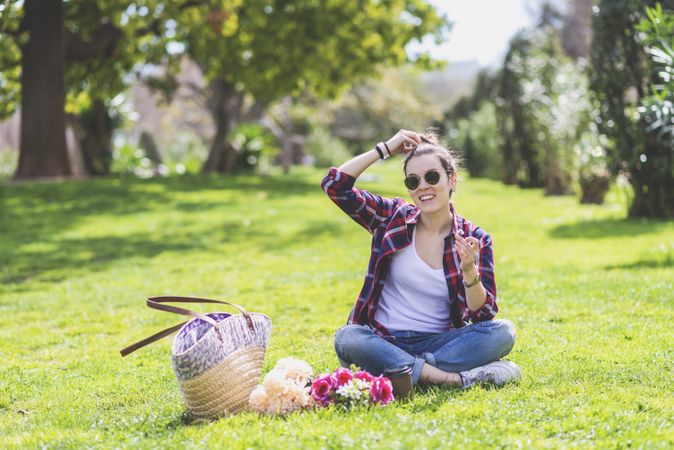 Front view of a young hipster woman sitting on grass in a park while holding a flower and smiling in a sunny day