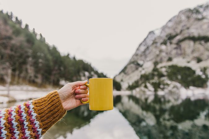 Person with cup near wonderful lake between hills in snow and cloudy sky