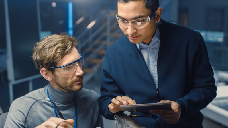 Two Professional Automotive Engineers with a Tablet Computer and Inspection Tools are Having a Conversation While Testing an Electric Engine in a High Tech Laboratory with a Concept Car Chassis.