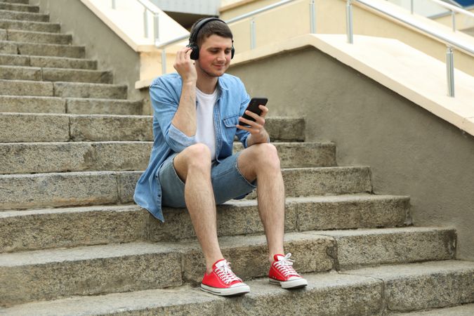 Young man in headphones sits on the steps and looking in phone