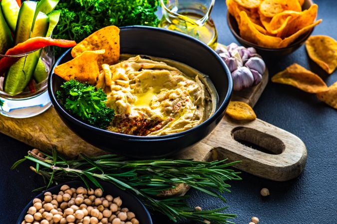 Creamy hummus dip in bowl on board served with olive oil, veggies and chick peas