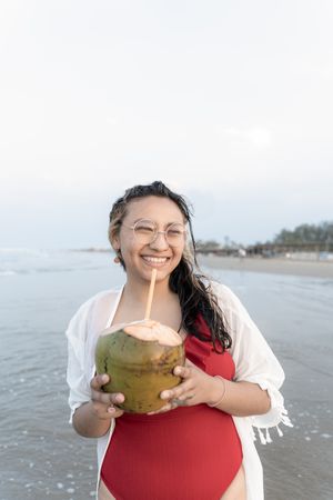 A young Hispanic woman is posing with a coconut on the sea edge at the beach