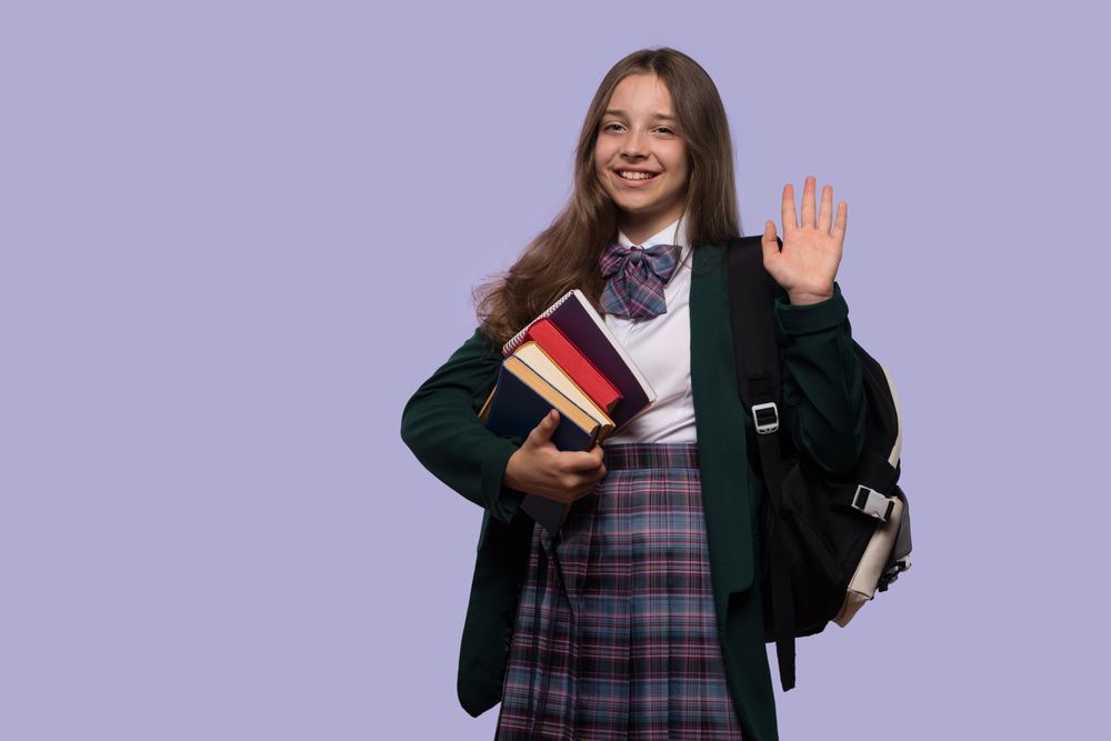 Smiling schoolgirl waves while holding stack of books.
