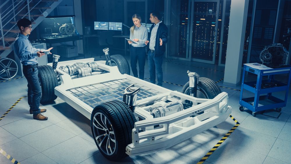 High Angle Shot of a Development Laboratory Room with Diverse Professional Team of Engineers and Scientists Working on a Electric Car Chassis with Wheels, Batteries, Engine and Suspension.