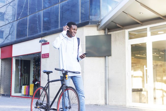 Smiling Black man walking in the street with bike while listening music on headphones