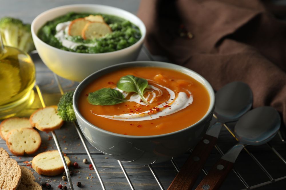 Two bowls of orange and green soup on wooden table with crackers