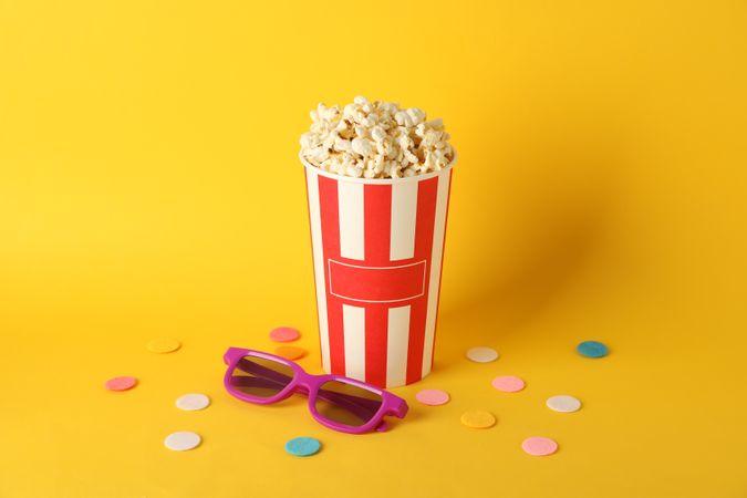 Striped bucket with popcorn and 3d glasses on yellow background, copy space