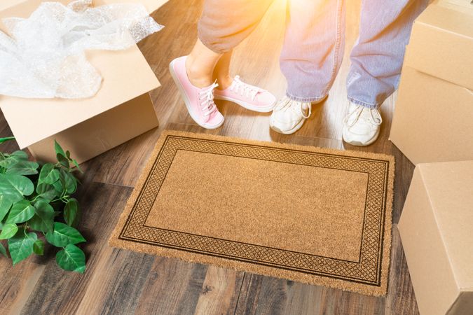 Man and Woman Standing Near Blank Welcome Mat, Moving Boxes and Plant