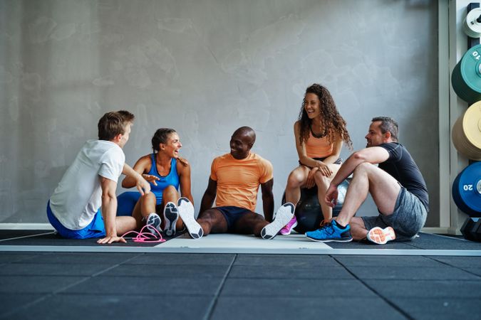 Multi-ethnic group of people in workout gear sitting on gym floor talking and laughing