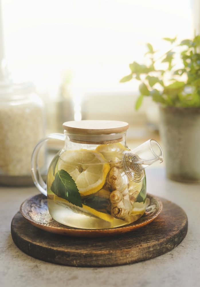 Glass teapot with lemon, mint and ginger on wooden cutting board at window background with natural light
