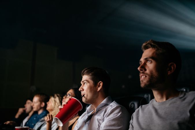 Young man having soft drink and watching movie with friends in cinema hall