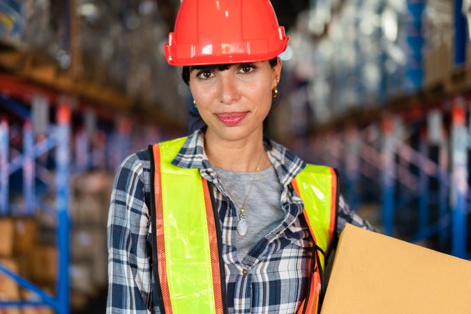 Smiling female warehouse worker in safety gear holding a box.