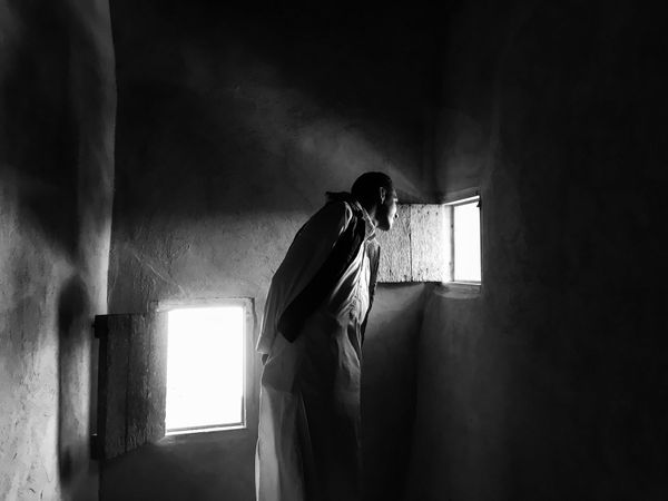 Grayscale photo of man in a small room looking through the small shutter in Siwa oasis, Egypt