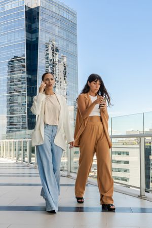 Two businesswomen are walking on a glass walkway. One of them is talking on the phone, and the other is sending a message.