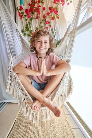 Smiling kid sitting on hammock chair and gesturing namaste