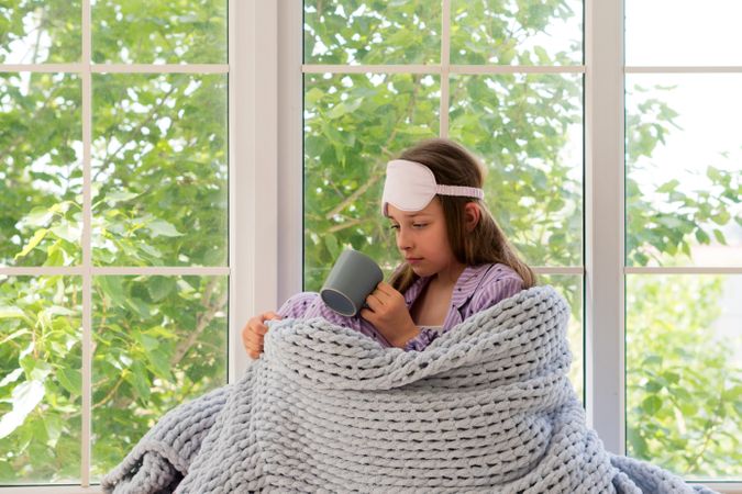 Teenage girl wrapped in grey knitted blanket by window.