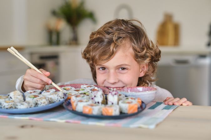 Happy boy hiding behind plates with sushi rolls in kitchen