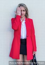 Portrait of a blonde woman wearing red jacket and skirt against a tiled wall covering her eye 48BNVZ