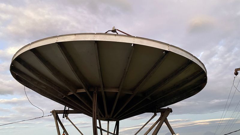 A large satellite dish stands prominently against a cloudy sky, its intricate structure exposed to the elements