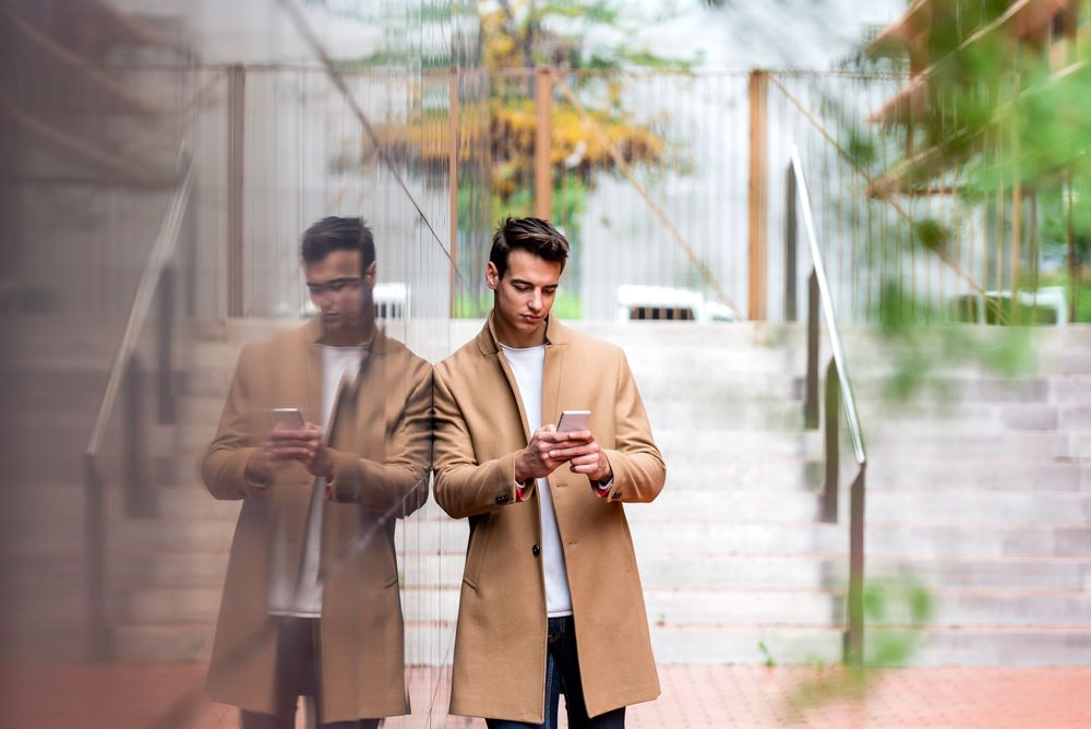 Man in fall coat leaning on reflective outdoor wall while texting
