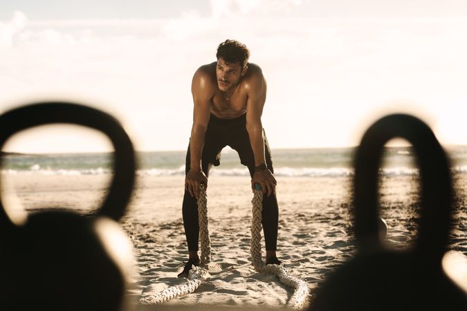 Exhausted man resting after fitness workout at a beach using battle ropes