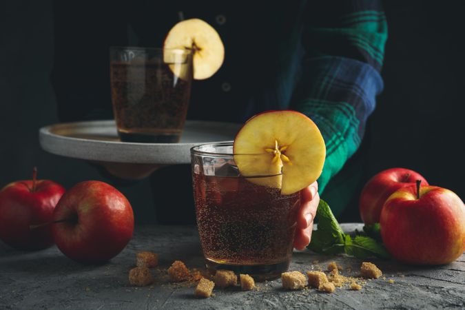 Female with tray hold glass of cider. Composition with cider and apple on gray table