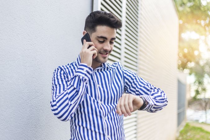 Young man standing outdoors and checking his wrist watch while speaking on phone