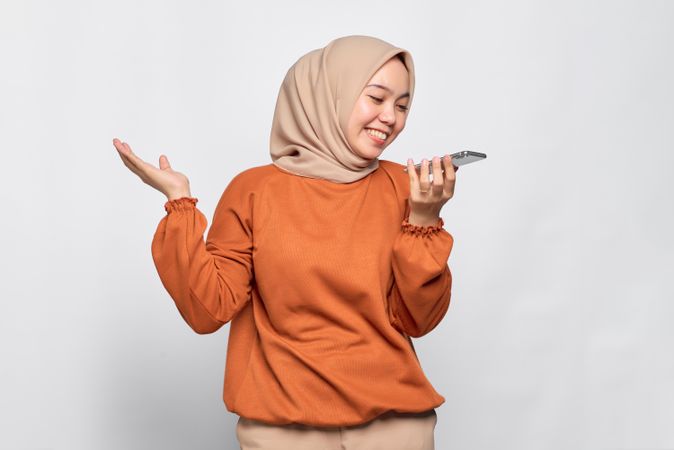 Smiling Muslim woman talking on smart phone speaker and making gesture with hand up