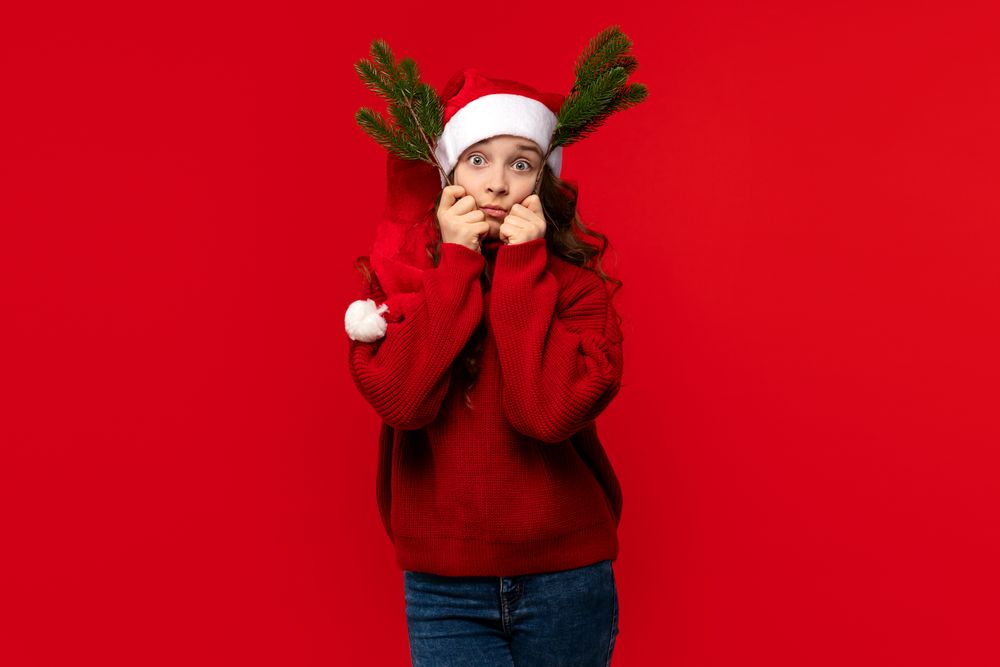 Girl in Santa hat making funny face with fir branches.
