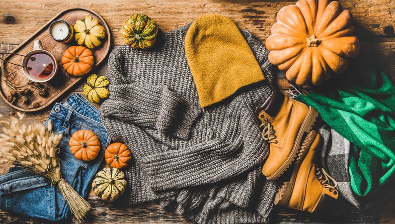 Flat-lay of grey sweater, blue jeans, green woolen scarf, hat, yellow boots, pumpkins and tea