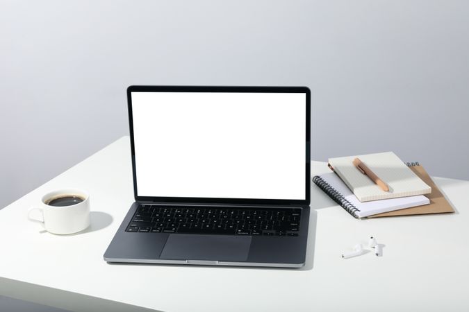 Table with open laptop with mockup screen, earbuds, coffee, and blank notebooks