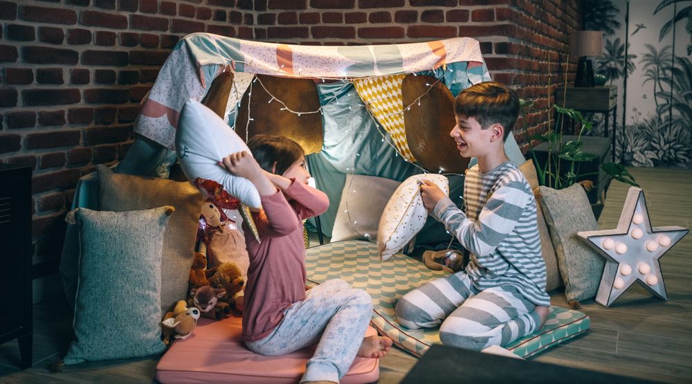 Funny children having fun playing pillow fight on handmade shelter tent in living room at home
