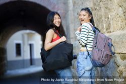 Two happy women in jeans outside looking back with bags and camera 5rr6n5
