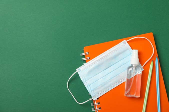 Medical mask, sanitizer, notebook and pencils on green background