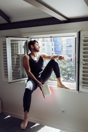Young male sitting on window and reading book