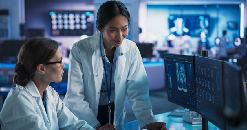 Medical Research Center Laboratory: White And Asian Female Scientists Talking And Using Desktop Computer With MRI Scans Of Human Brain.