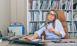 Blonde woman taking call, using computer and holding a nail file in office 4jVrkz