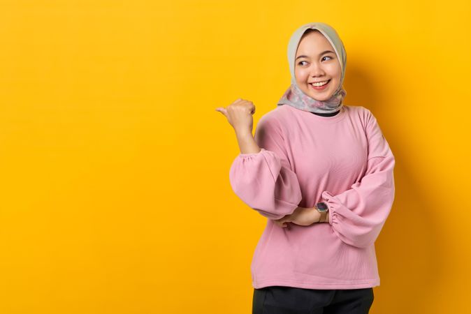 Smiling Muslim woman in scarf looking behind her and pointing with thumb