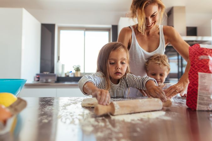Siblings with rolling pin and flour helping mother in kitchen at home