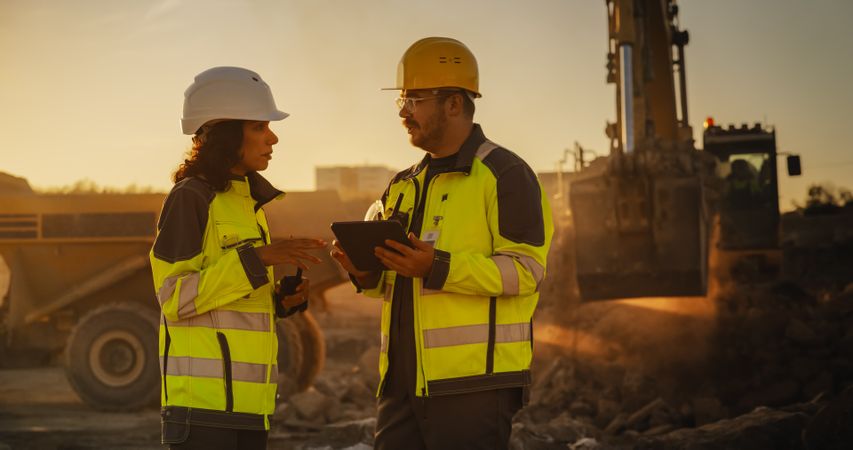 Cinematic Golden Hour Shot Of Construction Site: Male Civil Engineer And Hispanic Female Urban Planner Talking, Using Tablet. Trucks, Excavators, Loaders Working To Build New City District.
