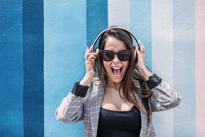 Excited woman wearing gray blazer and sunglasses listening to music on headphones