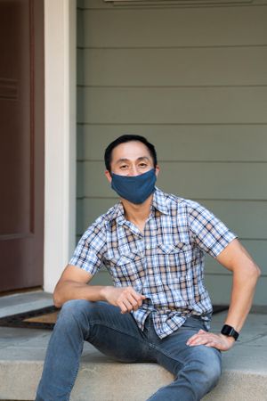 Man sitting in front of house wearing mask smiling and looking at camera
