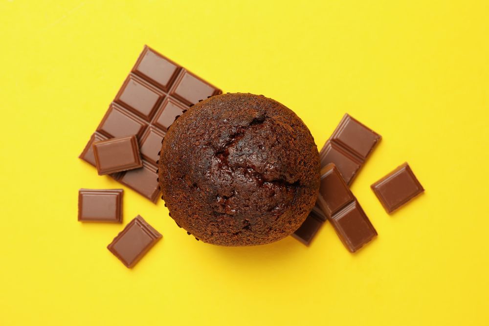 Tasty muffin and chocolate on yellow background, top view