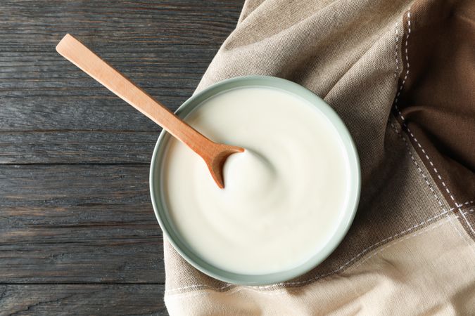 Bowl of sour cream yogurt, spoon and towel on wooden background