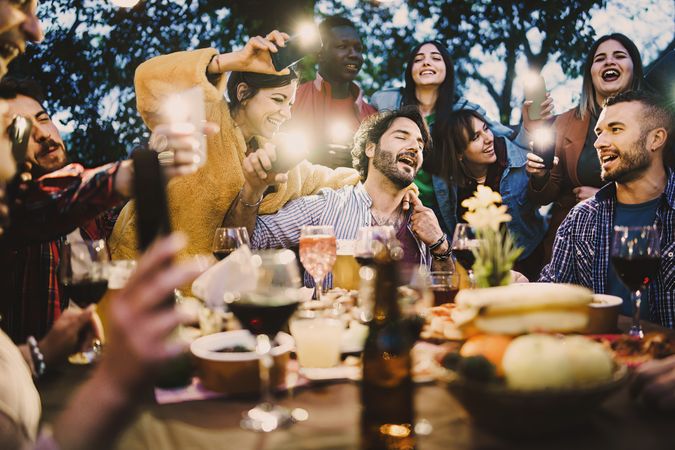 Group of drunk people singing around the barbecue food table drinking wine and beers and making lights with smart phones torches