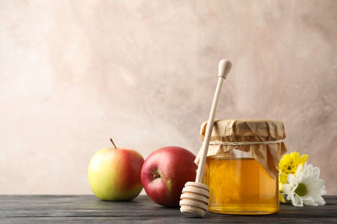 Glass jar with honey, dipper, apples and flowers on wooden background, space for text