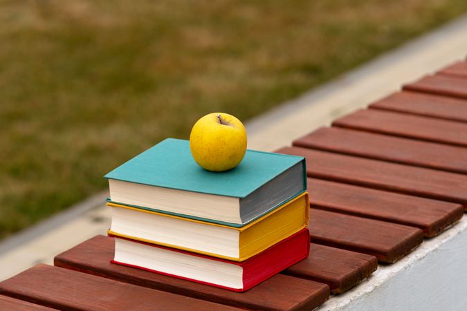 Stack of colorful hardcover books with apple on bench.