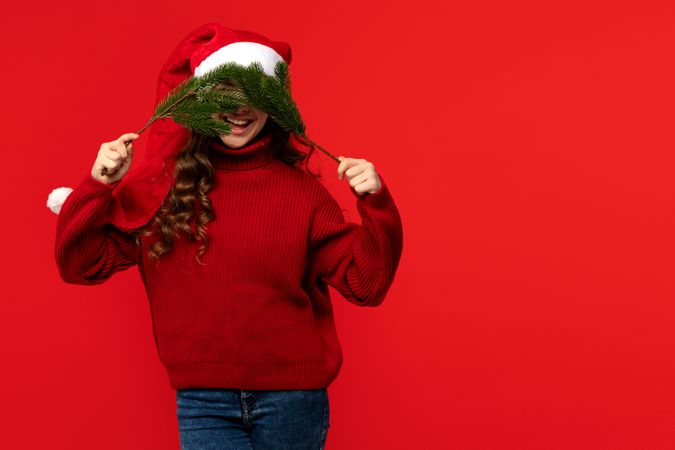 Girl in Santa hat playfully covering face with fir branches.