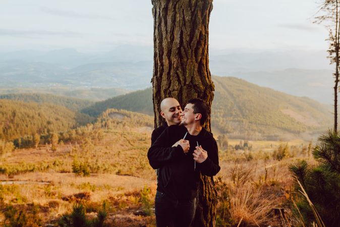 Smiling men hugging near tree in forest on hill